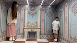 This is an Ames room illusion. The room is trapezoidal, not square, with a slanted floor and ceiling. Viewed from a specific angle, it appears normal, but as people move, perspective makes them seem to grow or shrink. It's an optical trick, not real size change. thumbnail